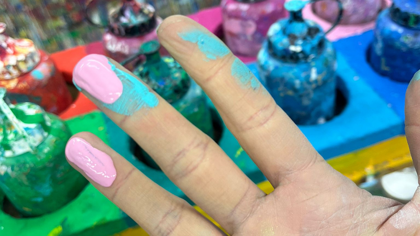Johannas Hand, die Fingerkuppen mit Farbe bedeckt. Im Hintergrund sieht man ein Holzaufbewahrung für verschiedene Farbflaschen der Firma Lascaux.