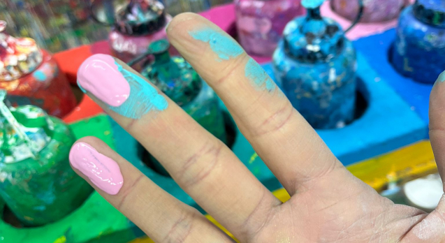 Johannas Hand, die Fingerkuppen mit Farbe bedeckt. Im Hintergrund sieht man ein Holzaufbewahrung für verschiedene Farbflaschen der Firma Lascaux.