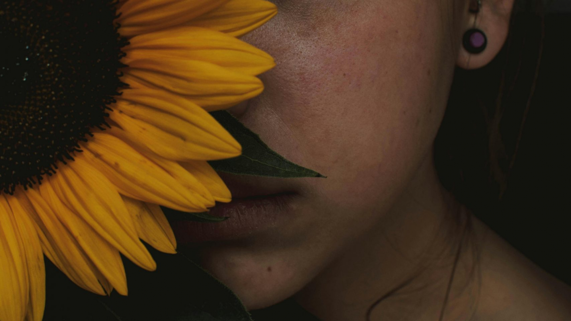 Frau mit Sonnenblume. Sie hält die Sonnen vor ihr Gesicht, sodass nur die linke Gesichtshälfte zu sehen ist. Das Bild verträumt und etwas dunkel. Die Frau blickt ernst, aber ihre Gesichtszüge sind entspannt