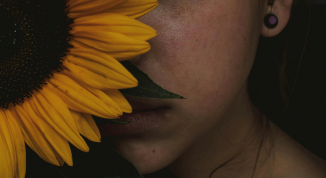 Frau mit Sonnenblume. Sie hält die Sonnen vor ihr Gesicht, sodass nur die linke Gesichtshälfte zu sehen ist. Das Bild verträumt und etwas dunkel. Die Frau blickt ernst, aber ihre Gesichtszüge sind entspannt